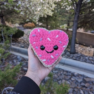 May include: A pink heart-shaped crocheted plush toy with white and purple sprinkles and a smiling face.