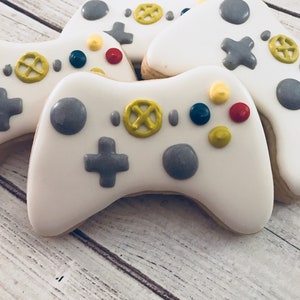 Peut inclure: Quatre biscuits en sucre décorés en blanc et gris en forme de manettes de jeu vidéo. Les biscuits ont des détails jaunes, rouges et bleus.