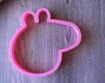 May include: A pink plastic cookie cutter in the shape of a pig's head. The cutter has a simple outline with rounded features, including ears and a snout. The cutter is on a wooden surface.