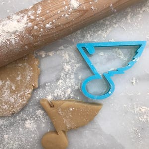 May include: A blue plastic cookie cutter shaped like a treble clef, with a rolled out piece of cookie dough in the shape of the treble clef next to it. A wooden rolling pin is in the background.