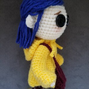 May include: Crocheted doll with blue hair, a yellow raincoat, and a burgundy bag. The doll is wearing yellow boots and has a red flower on her head.