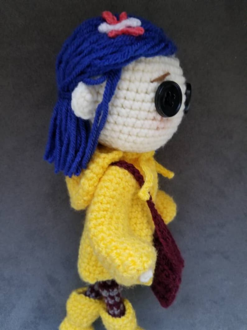 May include: Crocheted doll with blue hair, a yellow raincoat, and a burgundy bag. The doll is wearing yellow boots and has a red flower on her head.