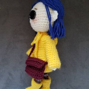 May include: Crocheted doll with blue hair, wearing a yellow raincoat, brown bag, and yellow rain boots.
