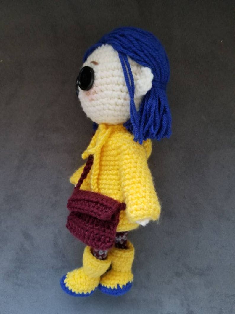 May include: Crocheted doll with blue hair, wearing a yellow raincoat, brown bag, and yellow rain boots.