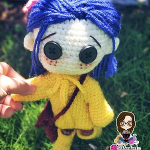 May include: Crocheted doll with blue hair, yellow sweater, brown bag, and big black button eyes. The doll is holding a small brown bag and is standing on a green grassy surface.