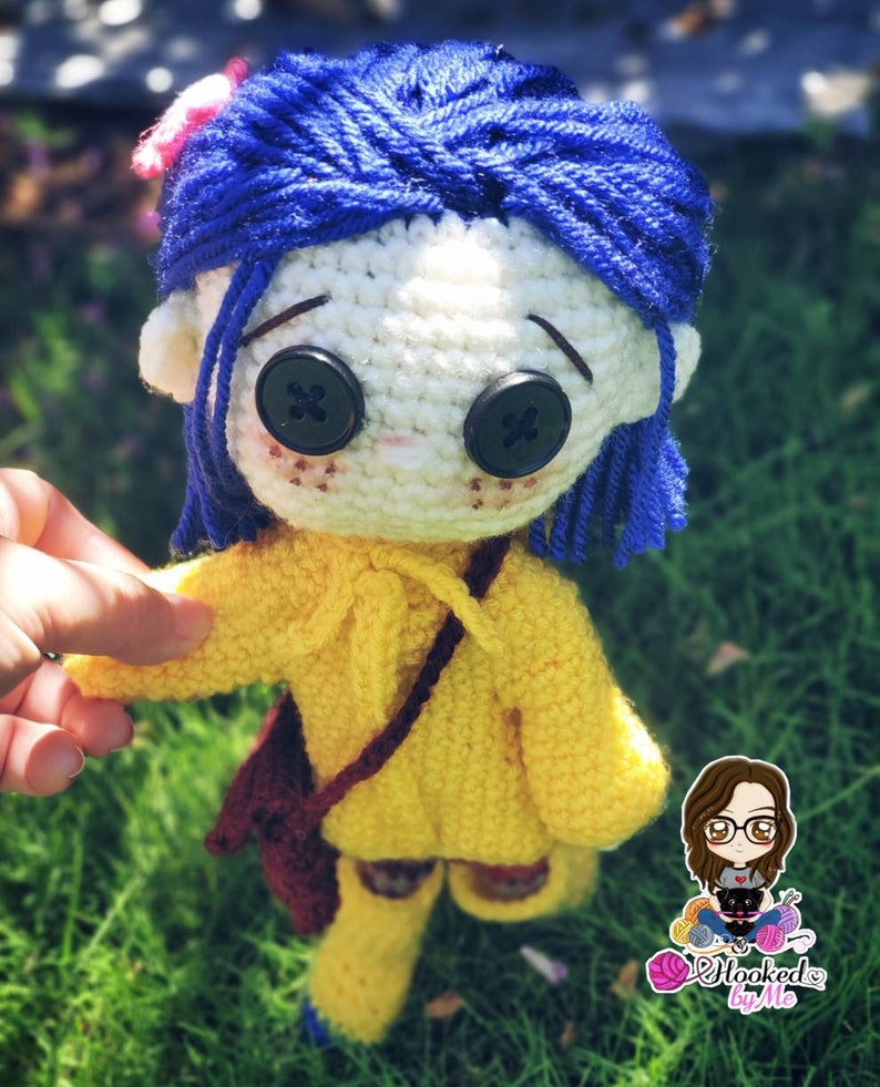 May include: Crocheted doll with blue hair, yellow sweater, brown bag, and big black button eyes. The doll is holding a small brown bag and is standing on a green grassy surface.