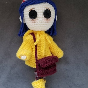 May include: A crocheted doll with blue hair, wearing a yellow raincoat, brown bag, and yellow boots with blue accents.
