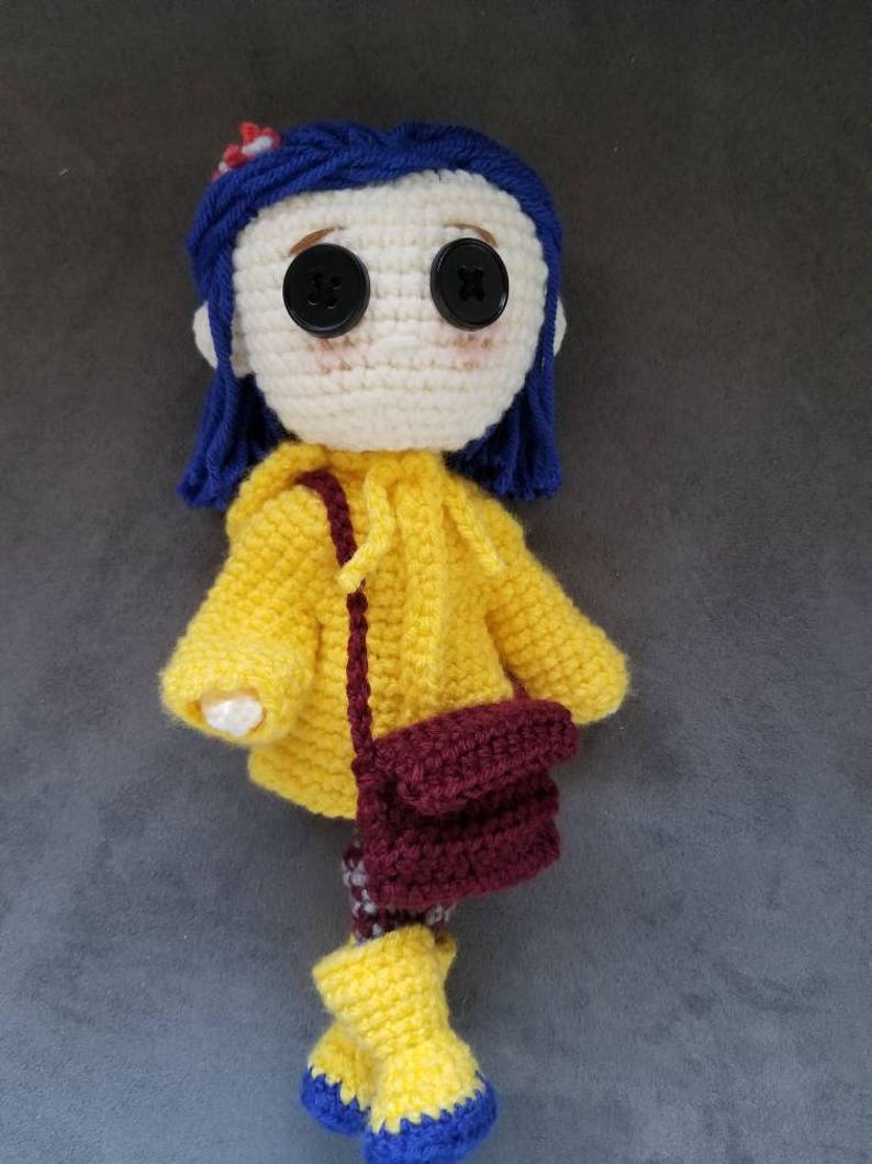May include: A crocheted doll with blue hair, wearing a yellow raincoat, brown bag, and yellow boots with blue accents.