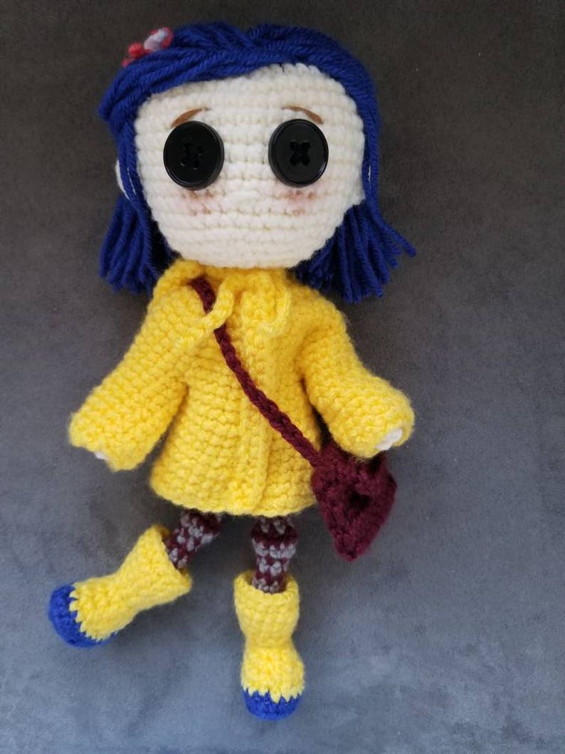 May include: Crocheted doll with blue hair, wearing a yellow raincoat, brown boots, and a burgundy bag.