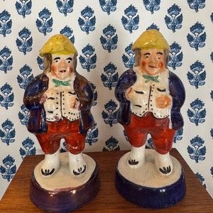 May include: Two ceramic figurines of men wearing blue jackets, red pants, and yellow hats. The figurines are standing on blue and white bases.