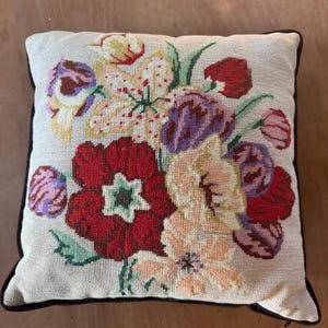 May include: Square decorative pillow with a floral needlepoint design. The pillow features a cream-colored background with a bouquet of red, purple, yellow, and pink flowers. The pillow has a dark brown border.
