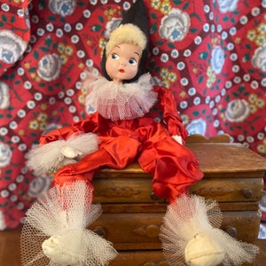 May include: A vintage doll dressed as a jester, wearing a red satin costume with white tulle ruffles and a black pointed hat. The doll is sitting on a wooden chest with drawers.