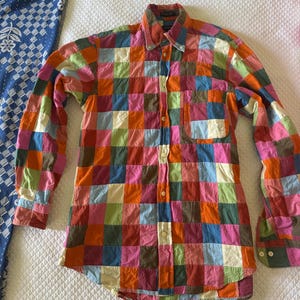 May include: A long-sleeve button-down shirt featuring a vibrant, multi-colored patchwork pattern of small squares in shades of orange, pink, blue, green, and yellow. The shirt includes a standard collar, a single chest pocket, and white buttons.