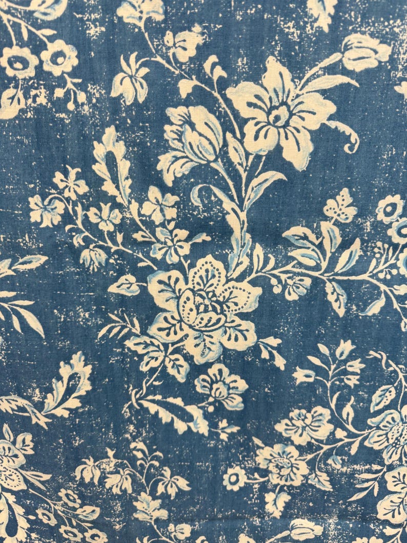 May include: Close-up of a blue fabric with a distressed floral pattern. The design features large, stylized flowers and foliage in off-white and light blue, creating a vintage aesthetic. The background is a deep blue with a textured, worn appearance.