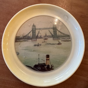 Könnte beinhalten: Ein runder, cremefarbener Untersetzer mit einer detaillierten Illustration der Tower Bridge in London. Das Bild zeigt Boote auf dem Wasser und ein Schleppboot im Vordergrund. Der Untersetzer hat einen leicht erhöhten Rand.