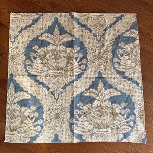 May include: A square fabric piece with a repeating pattern of floral and ornamental designs in blue, white, and beige. The fabric features a central floral motif within a diamond-shaped frame.