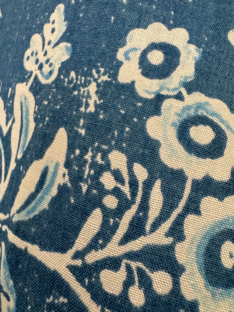 May include: Close-up of a blue and white floral pattern on fabric. The design features stylized flowers and leaves in shades of blue and white against a textured blue background. The fabric appears to be cotton or a similar material.
