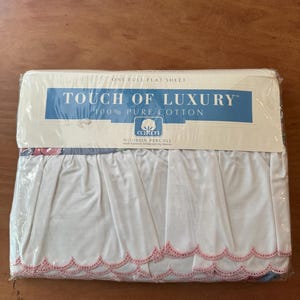May include: A white flat sheet with a pink scalloped edge. The packaging says "Touch of Luxury" and "100% Pure Cotton" with a cotton boll logo. It also says "No-Iron Percale".