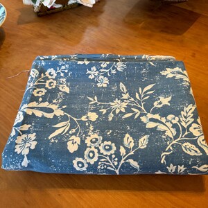 May include: Folded blue fabric with a white floral pattern. The fabric features a distressed, vintage-inspired design with flowers and leaves. The fabric is folded neatly on a wooden surface.