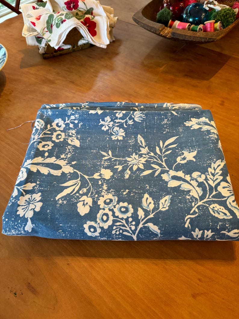 May include: Folded blue fabric with a white floral pattern. The fabric features a distressed, vintage-inspired design with flowers and leaves. The fabric is folded neatly on a wooden surface.