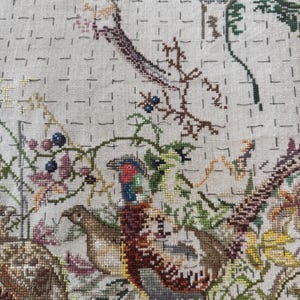 May include: A detailed cross-stitch embroidery featuring pheasants and other birds amidst foliage. The artwork showcases a variety of colors, including greens, browns, and blues, with a textured, hand-stitched appearance. The design includes berries and branches.
