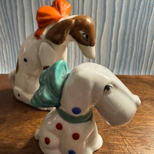 May include: Two ceramic dog figurines. One dog has an orange bow, brown ears, and blue spots. The other dog has a teal bow, green collar, red and blue spots. Both dogs are white with a glossy finish.