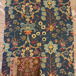 May include: A rectangular piece of fabric featuring a dark blue background with a repeating pattern of stylized yellow, red, and light green floral motifs. A smaller, square patch of brown fabric with a similar floral print is attached at the bottom.