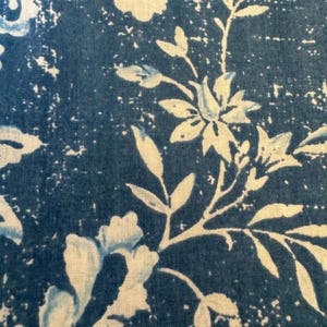 May include: Close-up of a fabric with a blue and white floral pattern. The design features stylized flowers and leaves against a textured, dark blue background. The white elements have a slightly distressed appearance.