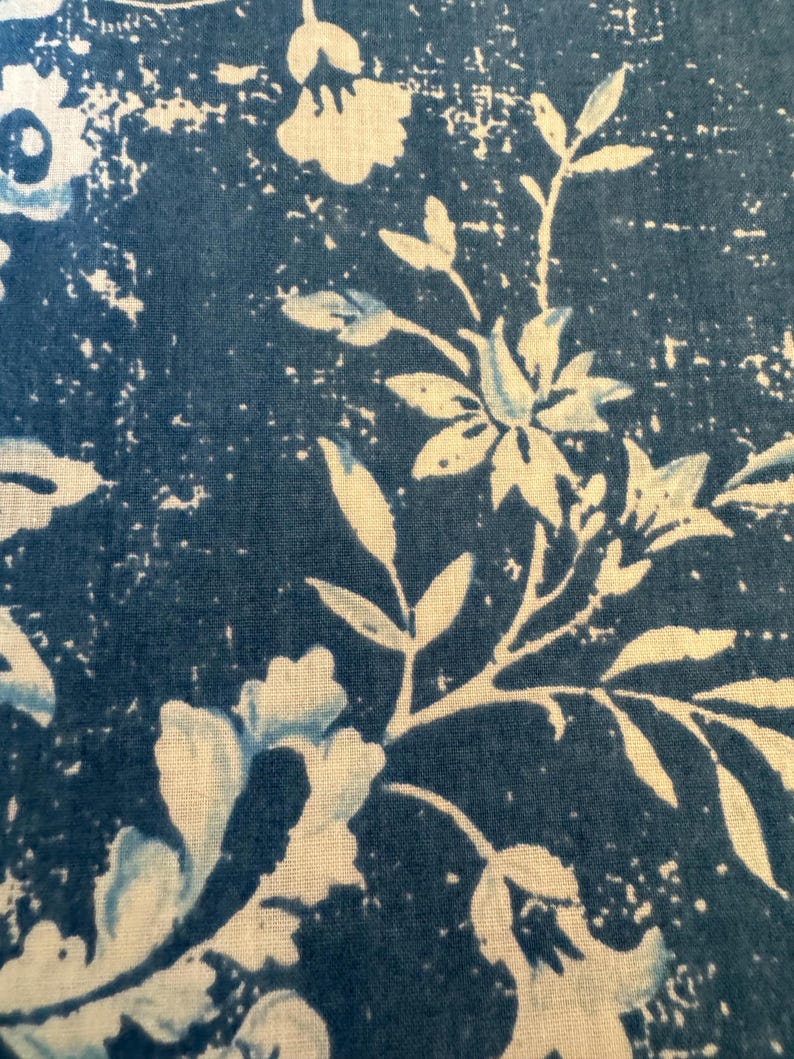 May include: Close-up of a fabric with a blue and white floral pattern. The design features stylized flowers and leaves against a textured, dark blue background. The white elements have a slightly distressed appearance.