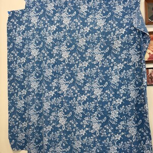 May include: A blue floral fabric with a white flower pattern is displayed. The fabric is hanging on a wall, held in place with blue tape. The floral design is detailed and covers the entire surface of the fabric.