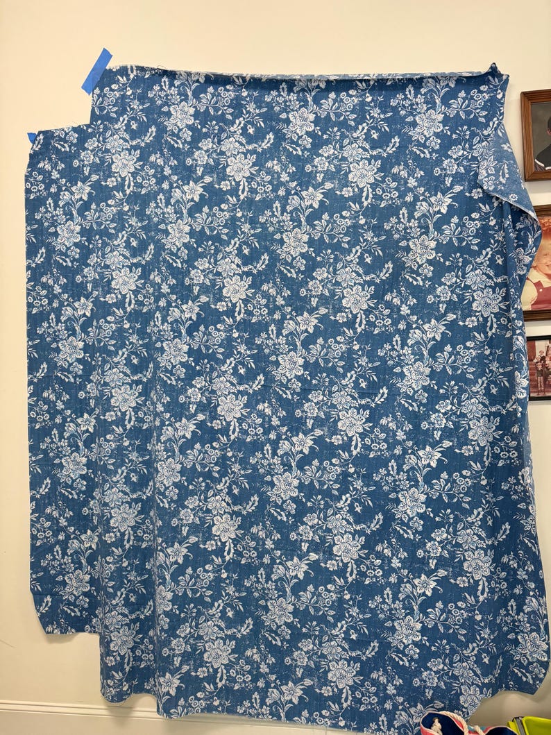 May include: A blue floral fabric with a white flower pattern is displayed. The fabric is hanging on a wall, held in place with blue tape. The floral design is detailed and covers the entire surface of the fabric.