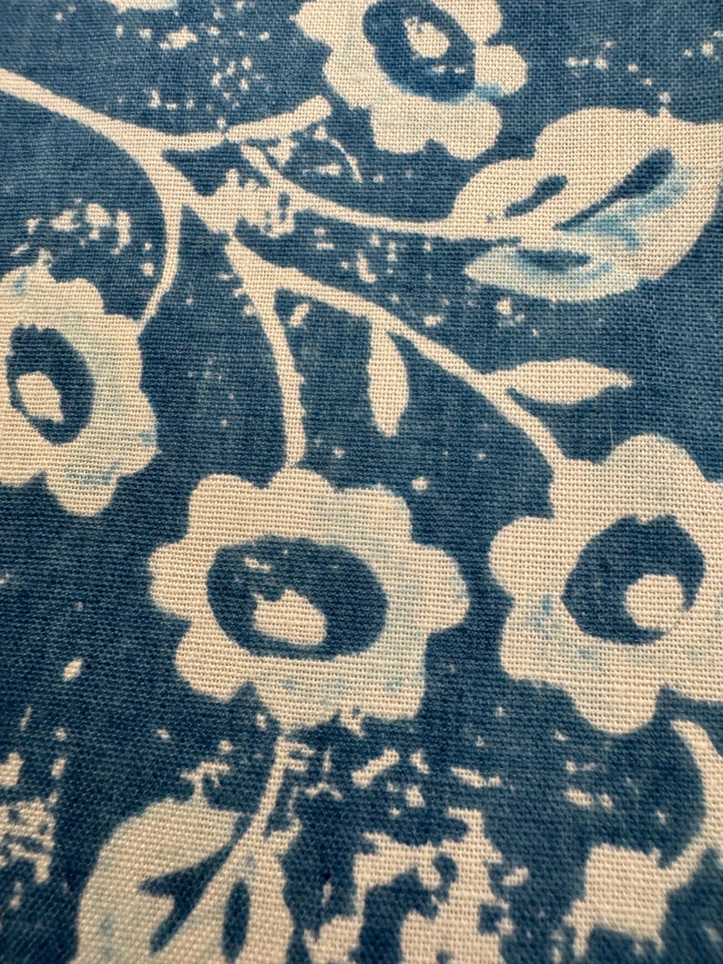 May include: Close-up of a fabric with a repeating floral pattern. The design features white flowers and leaves against a deep blue background. The fabric has a textured appearance, with visible weave details.