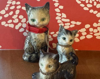 May include: Three ceramic cat figurines, each with black and white markings and green eyes. The largest cat is wearing a red collar with a gold chain attached. The other two cats are wearing gold chains around their necks.