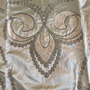 May include: A close-up of a beige fabric with a decorative pattern of a stylized fleur-de-lis design in a light brown color. The pattern is outlined in a light blue color and features a series of small circles.
