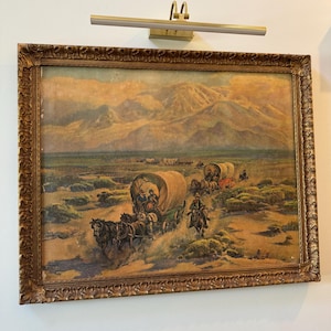 May include: An antique framed painting depicting a Western scene with covered wagons, horses, and riders traversing a desert landscape. The artwork features a detailed, ornate gold frame and is mounted on a white wall. A brass picture light is mounted above the painting.