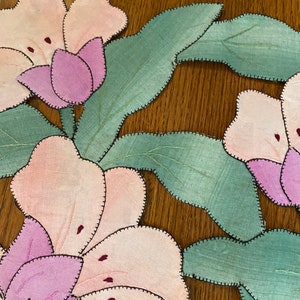 May include: A close-up of a handmade fabric applique featuring pink and purple flowers with green leaves. The fabric is stitched together with a black thread, creating a decorative design.