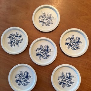 Könnte beinhalten: Sechs weiße Untersetzer mit einem blauen Hummer-, Bier- und Stechpalmen-Design. Die Untersetzer sind rund und haben einen leicht erhöhten Rand. Das Design hat ein klassisches nautisches Thema.