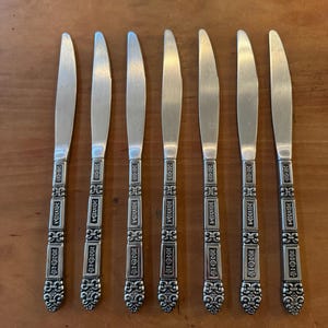 May include: Seven silver butter knives with ornate handles are arranged on a wooden surface. The knives have a simple blade design and decorative, detailed handles. The handles feature a repeating pattern of rectangular and floral elements.