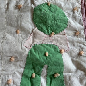 May include: A handmade quilt featuring a whimsical figure with a green hat, shirt, and pants. The figure is appliquéd onto a white background, with small, fluffy, beige embellishments scattered around. The quilt has a pink border and is likely a children's item.