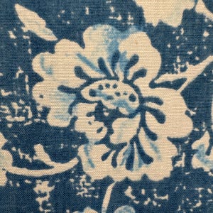 May include: Close-up of a blue and white floral pattern. The design features large, stylized flowers with intricate details, set against a textured blue background. The fabric appears to be cotton or a similar material, with a slightly faded or vintage aesthetic.