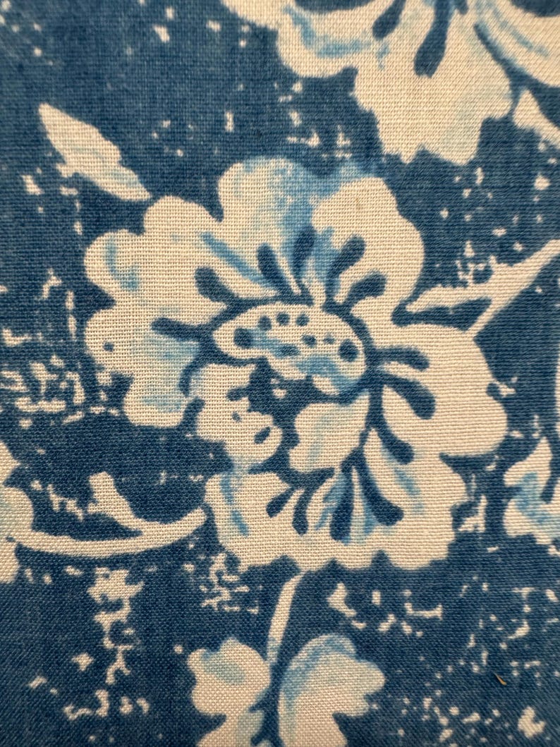 May include: Close-up of a blue and white floral pattern. The design features large, stylized flowers with intricate details, set against a textured blue background. The fabric appears to be cotton or a similar material, with a slightly faded or vintage aesthetic.