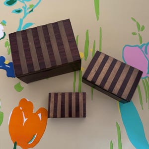 May include: Three wooden boxes with a striped pattern. The boxes are different sizes and are made of dark wood with gold stripes.