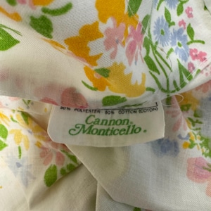 May include: Close-up of a white fabric with a floral pattern in yellow, pink, and blue. The fabric has a tag that reads "50% Polyester 50% Cotton (Coton) Cannon Monticello."