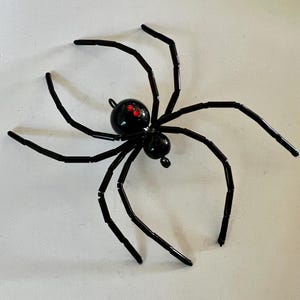 Puede incluir: Decoración de araña negra con ocho patas articuladas y un cuerpo redondeado. El cuerpo de la araña tiene dos segmentos, con detalles rojos para los ojos. Esta decoración de Halloween está hecha de un material brillante.