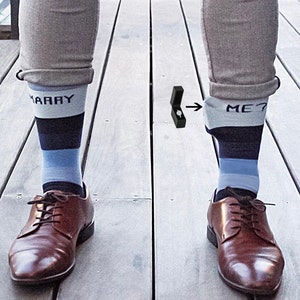 May include: Brown leather dress shoes paired with socks that say "MARRY ME?" in blue and white. A black ring box with a diamond ring is next to the socks. The socks have blue and navy stripes.