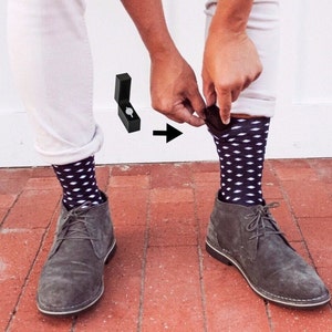 May include: A man is hiding a small black ring box in his sock. The ring box is visible in the image. The man is wearing white pants, grey suede shoes, and black and white polka dot socks.