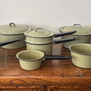 May include: A collection of vintage olive green enamelware pots and pans with black handles. The set includes various sizes of pots, saucepans, and a double boiler, all with black rims. The cookware is arranged on a wooden surface.