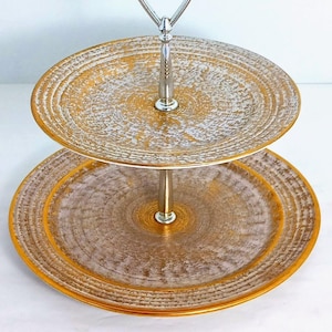 May include: Two-tiered serving stand with gold and white distressed finish. The plates have a textured, circular design with a gold rim. The handle and central support are silver-toned. Ideal for displaying desserts or appetizers.