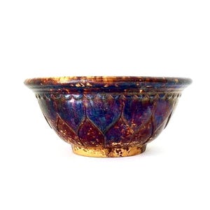 May include: A ceramic bowl with a glossy, iridescent finish. The bowl features a deep purple and blue color scheme with a gold-colored base. The exterior is decorated with a repeating petal pattern.