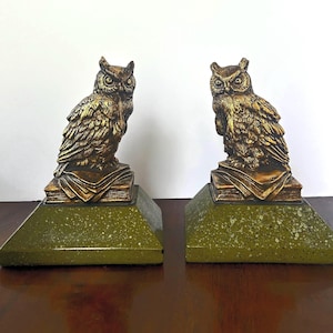 May include: Pair of owl bookends. Each bookend features a detailed gold-colored owl perched atop a stack of books. The base is a green, speckled, trapezoid shape. The bookends are made of a metallic material.